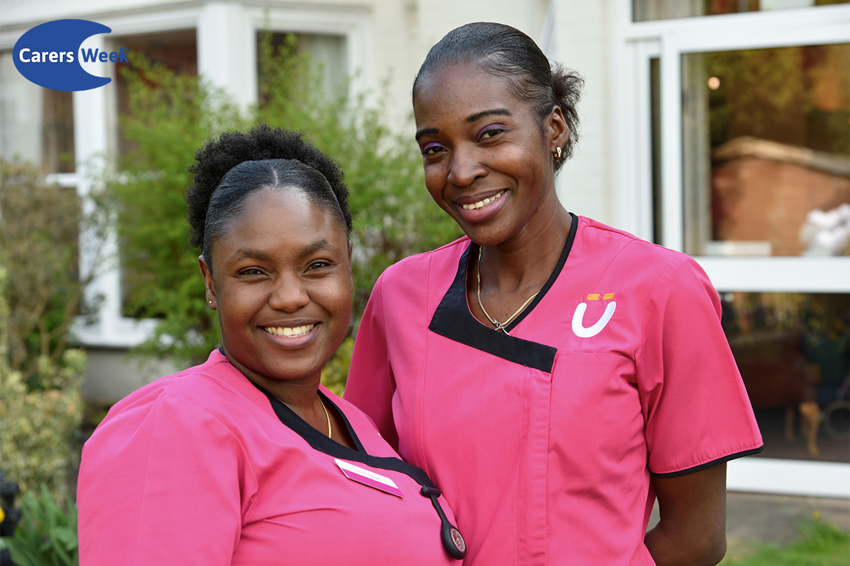 Two Care Plus carers in their pink uniforms. They are standing outside in front of foliage and a building.